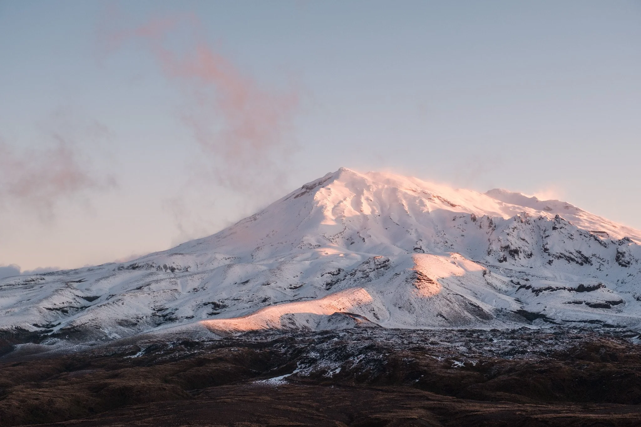 A stunning photo by Lou Lou B Photo of the vibrant pink after-sunset colors in the sky above Mount Ruapehu during winter. The snow-capped peak is silhouetted against the sky, with wispy pink clouds appearing to radiate from the mountain's summit. Key