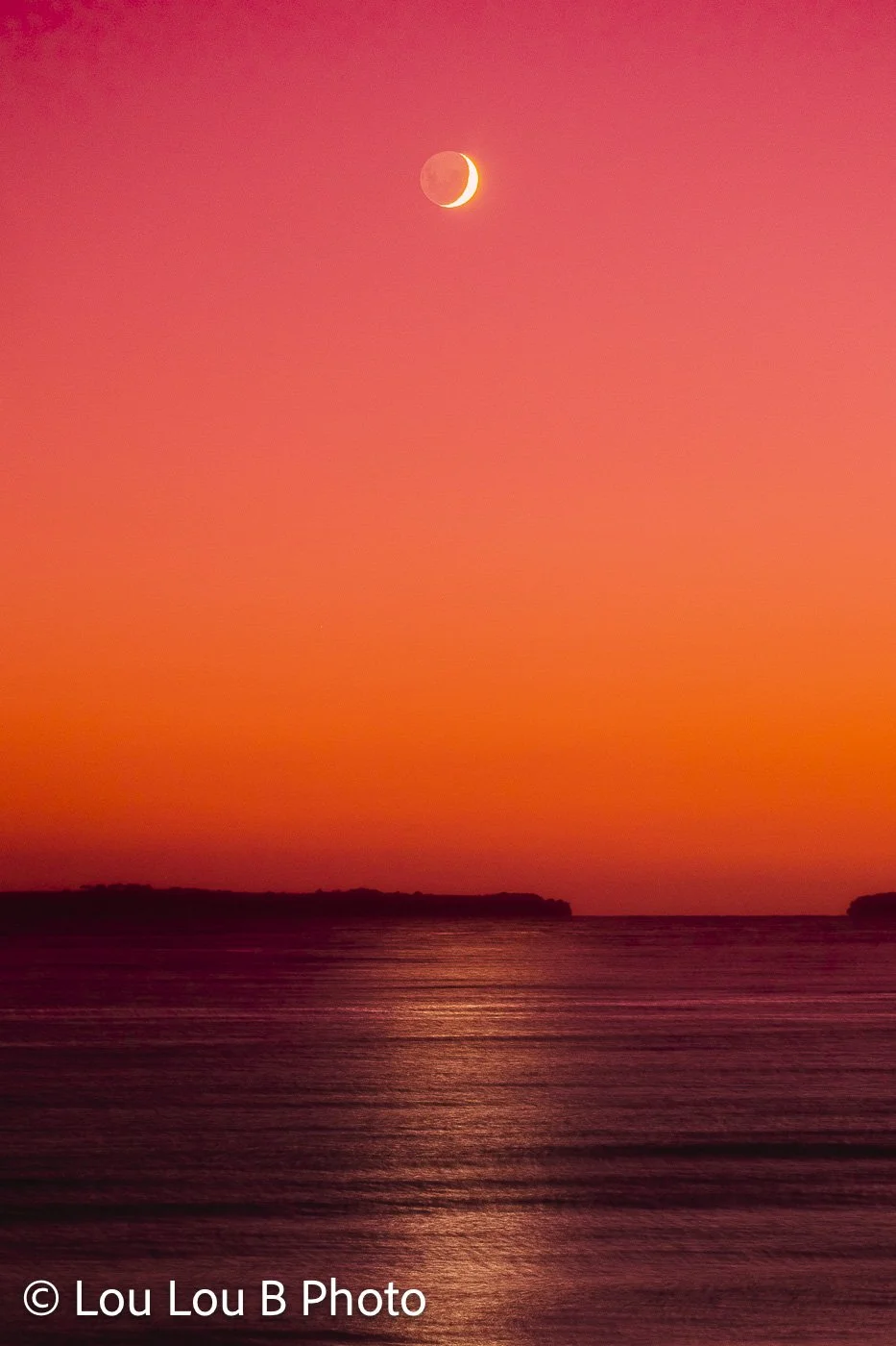 Crescent moon above the ocean in a vivid red and orange sunset sky, with light reflected across the water.
