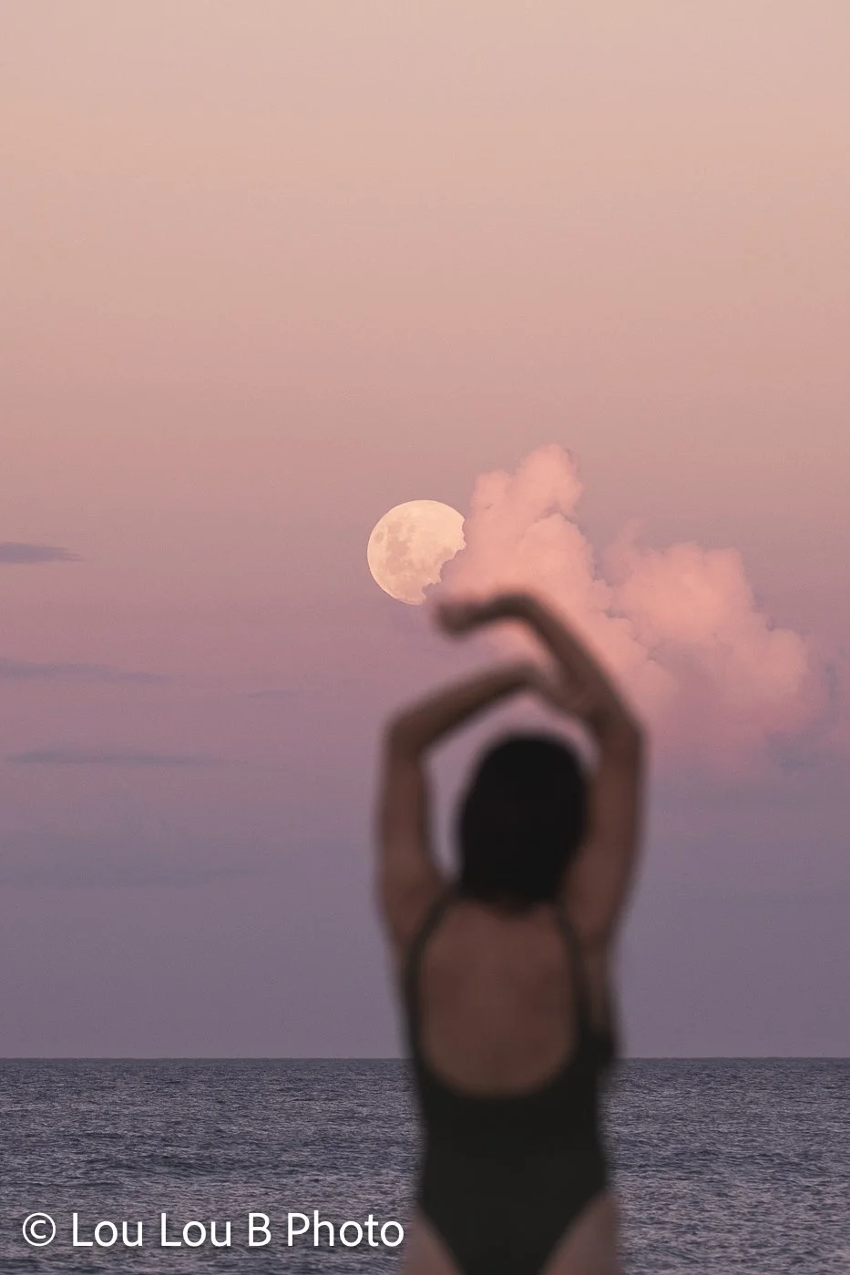 Woman silhouetted beneath a full moon and pink cloud, with her arms raised above her head at the beach.