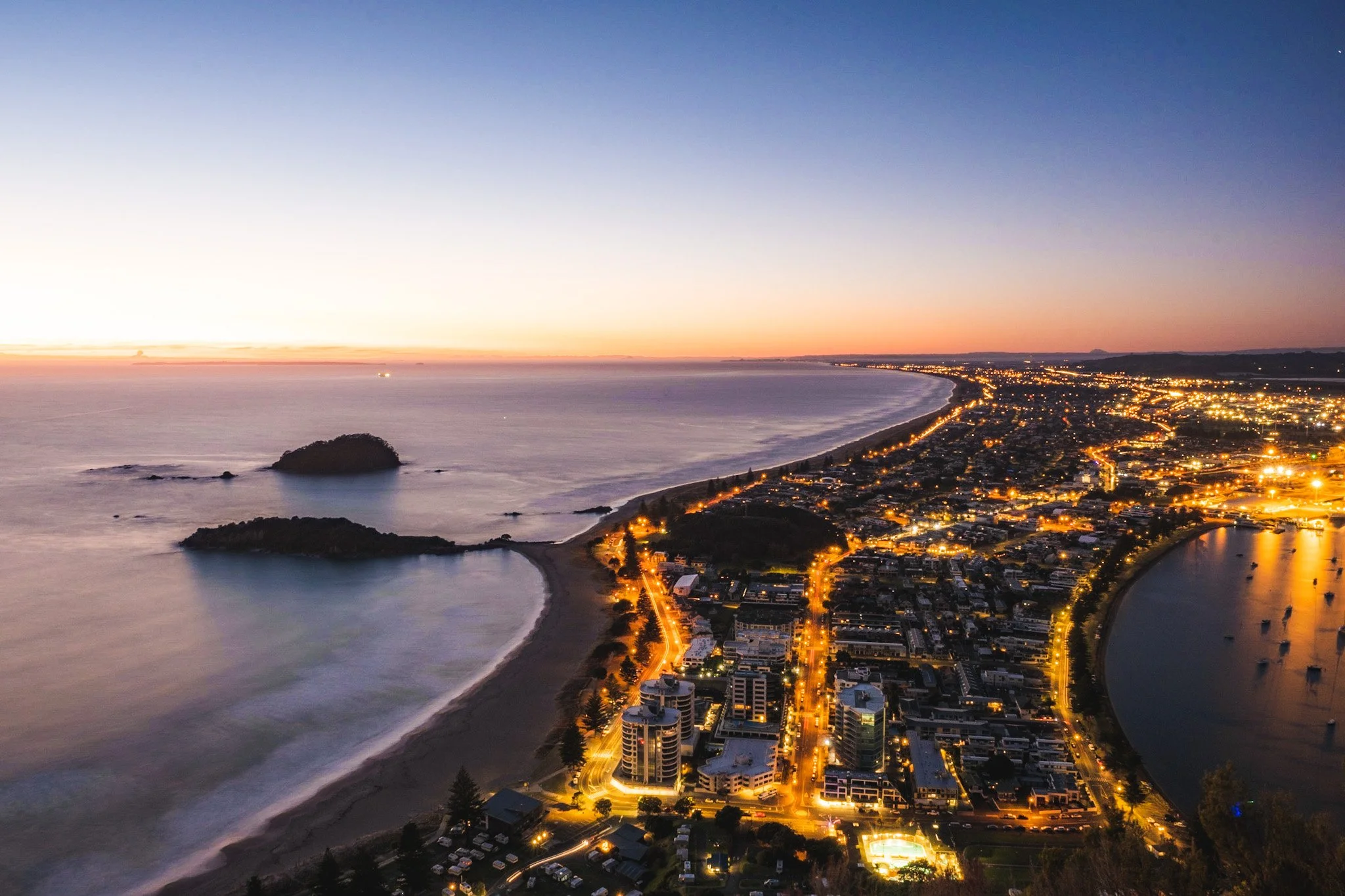 Experience the stunning view from the top of Mount Maunganui with this captivating photograph, taken just before sunrise by Lou Lou B Photo. The ocean below is captured in motion with a slow shutter, and the lights of the town twinkle in the distance