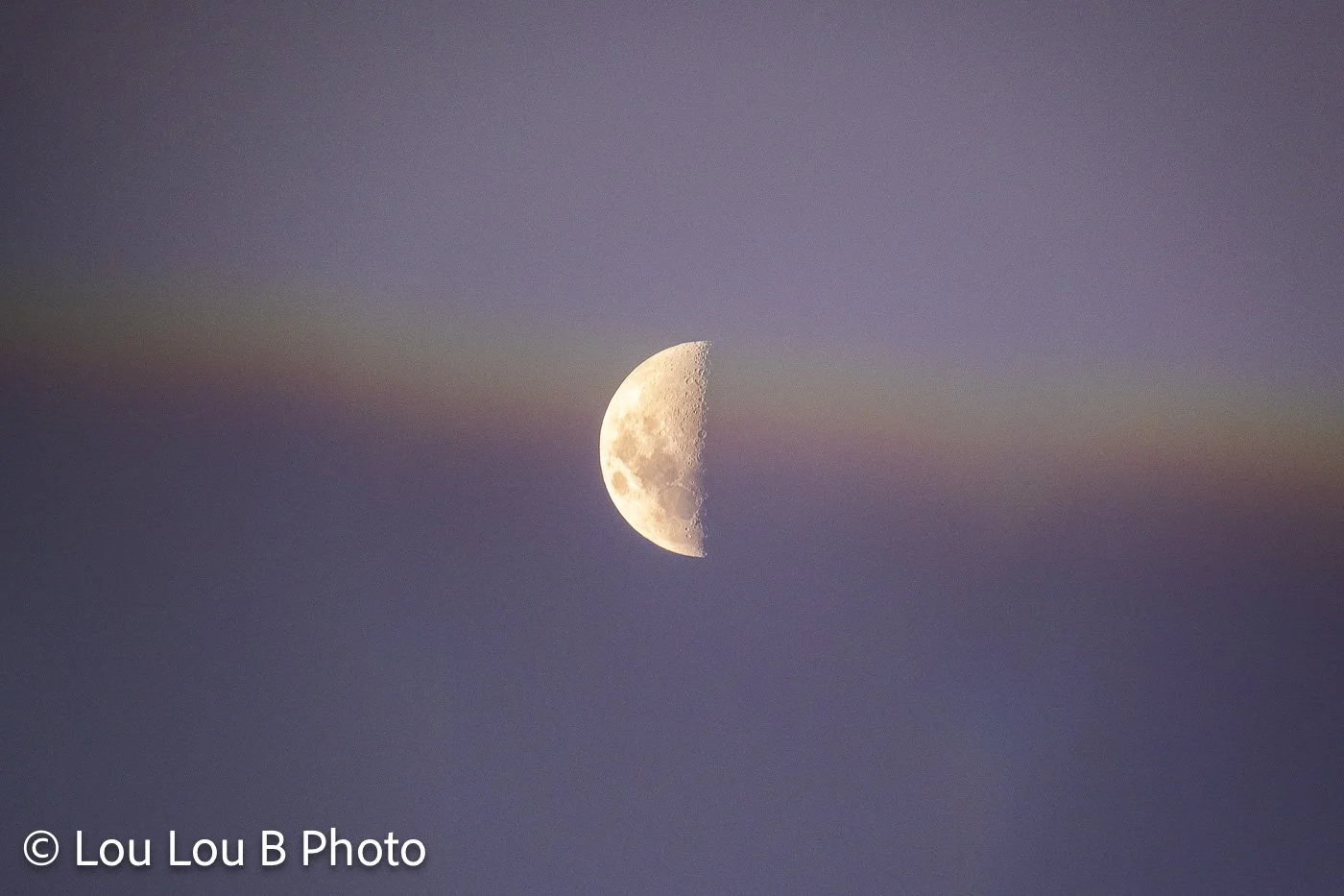 Half moon emerging through a soft band of atmospheric rainbow cloud in a lavender-blue sky.
