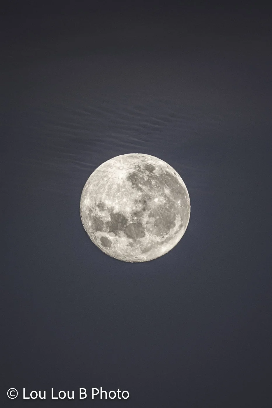 Detailed pale full moon rising through a hazy blue evening sky.