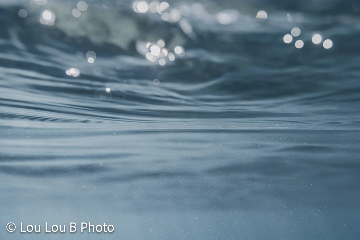 Close-up of a translucent blue ocean wave with sparkling sunlight across the water.