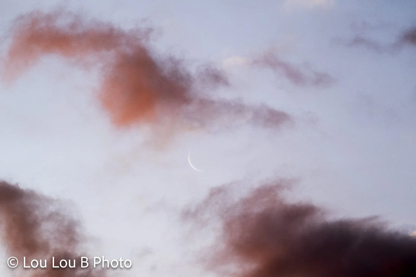 Delicate crescent moon emerging between pink and purple clouds in a pale blue twilight sky.