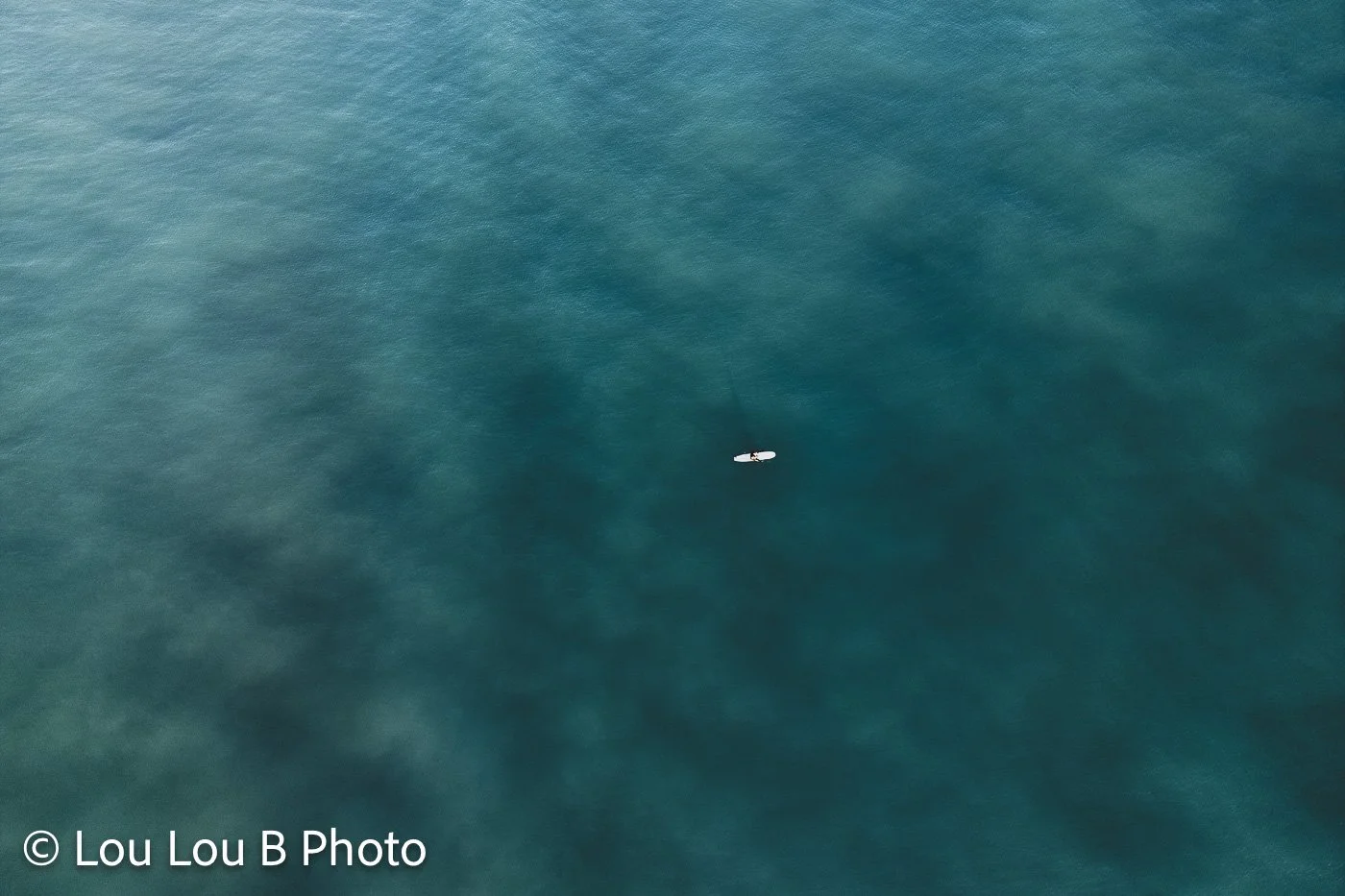 Aerial view of a lone surfer in deep turquoise ocean water.