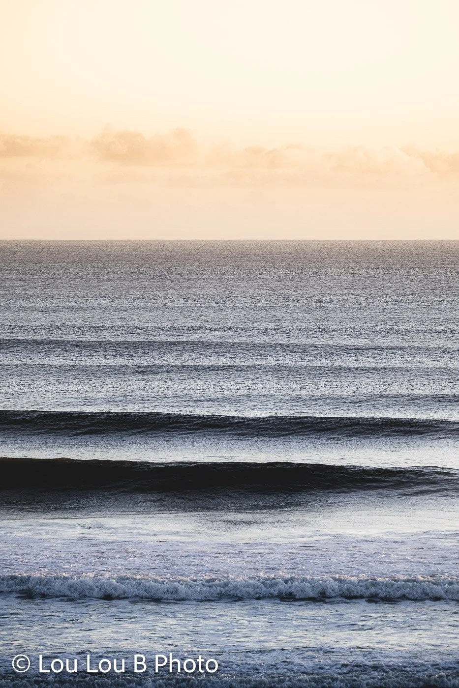 Parallel ocean waves rolling toward shore beneath a soft champagne and pale gold sky.