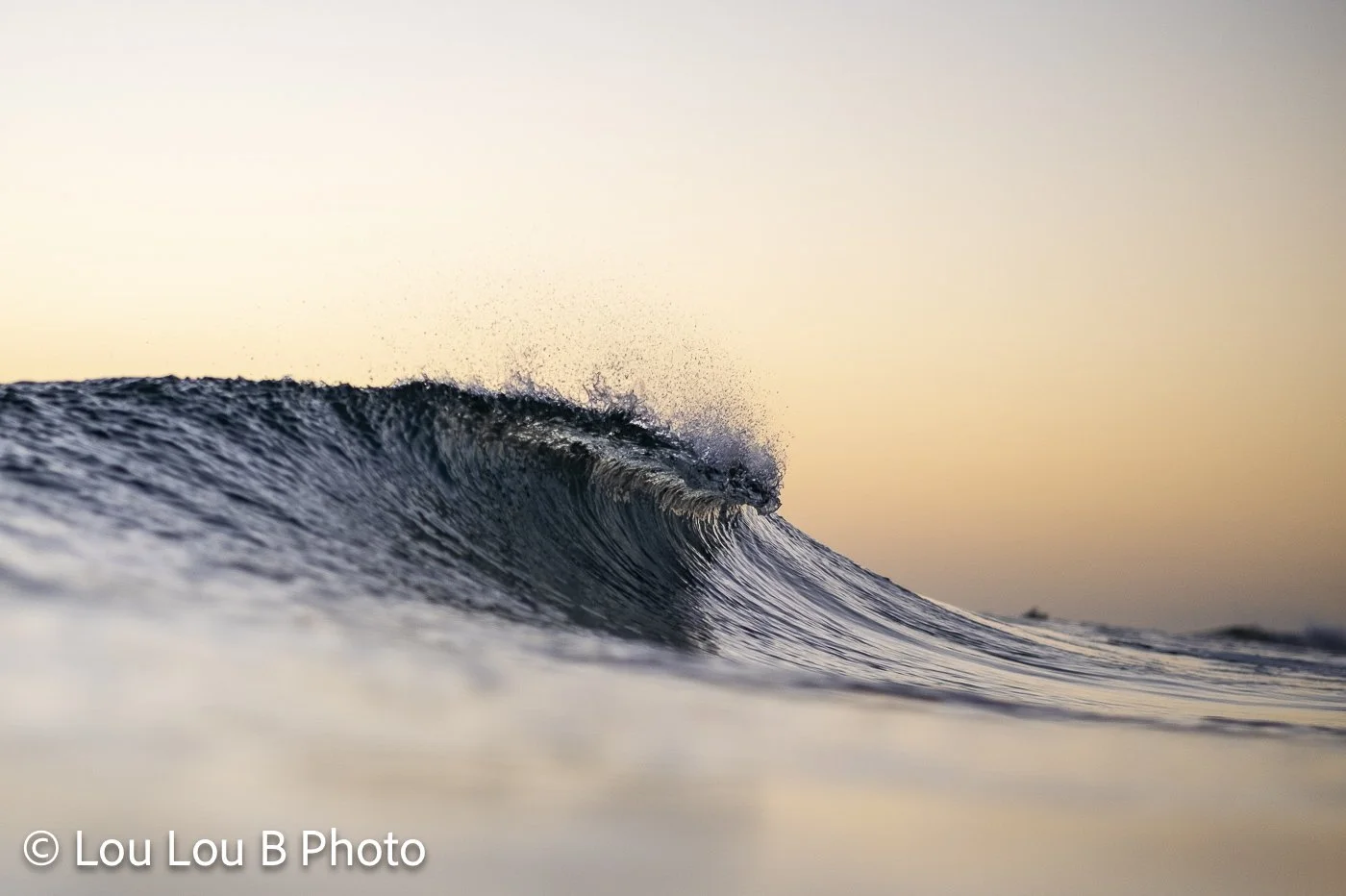 Close-up of a curling blue wave against a soft peach and champagne-coloured sky.