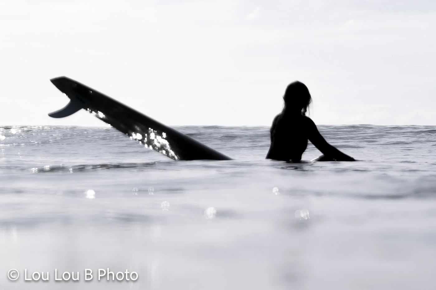 Silhouette of a surfer standing in calm silver-blue water beside a surfboard.