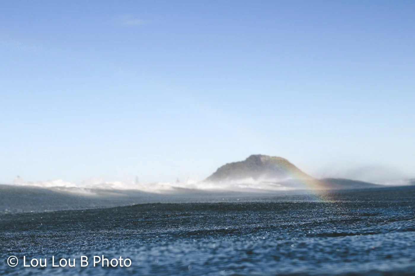 Mauao through sea spray with a rainbow photographed from the water at Mount Maunganui