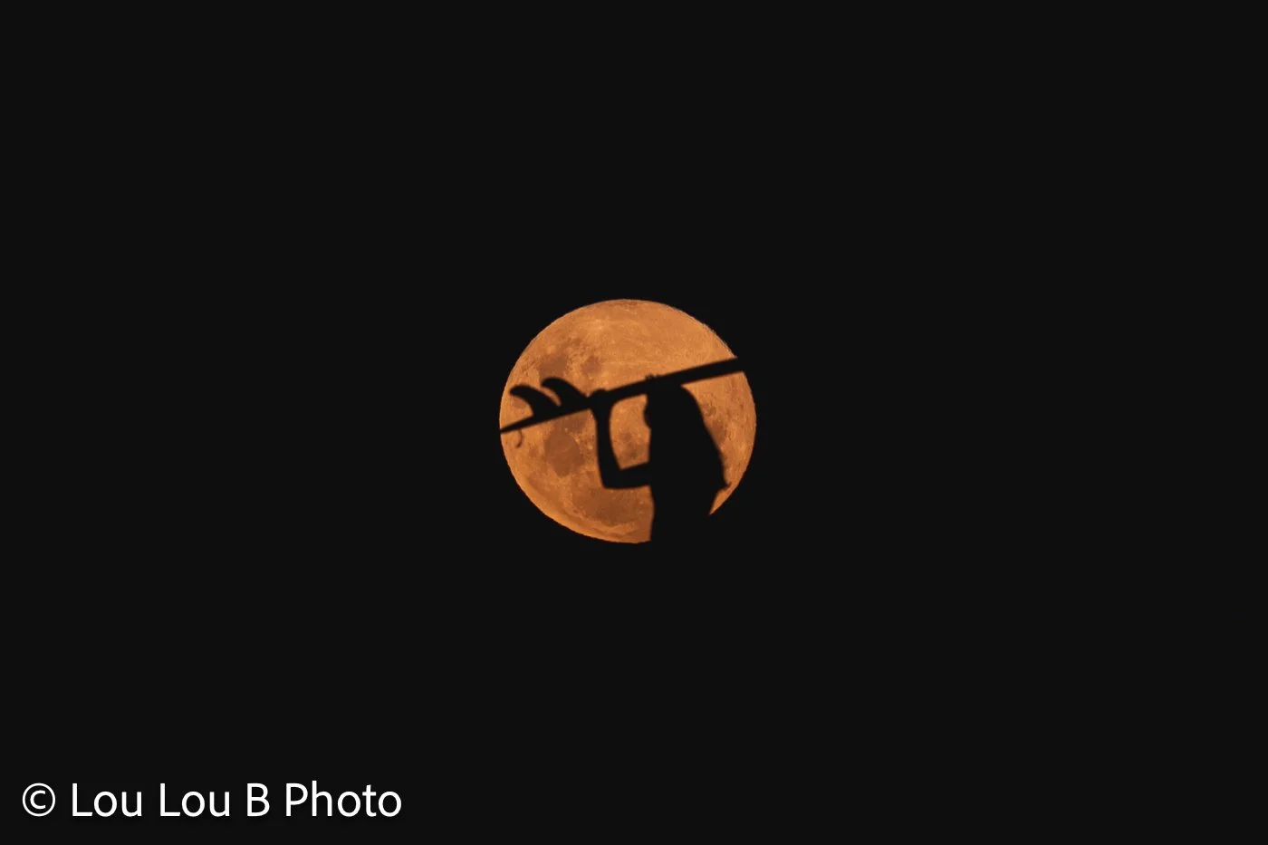 Silhouette of a woman carrying a surfboard across a large orange full moon.