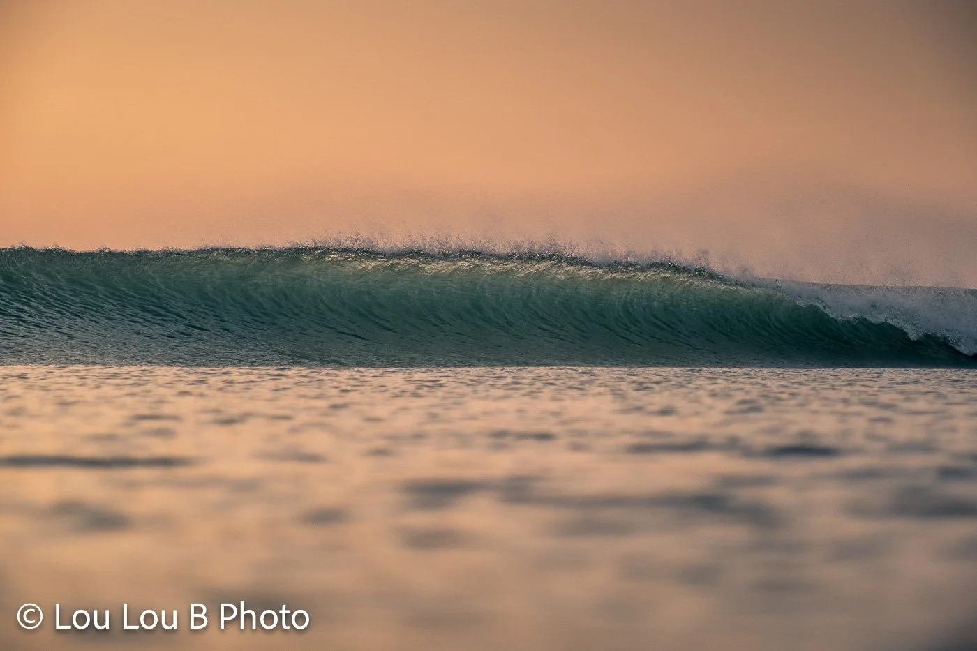 Long turquoise wave breaking beneath a warm peach sky with fine spray lifting from the crest.
