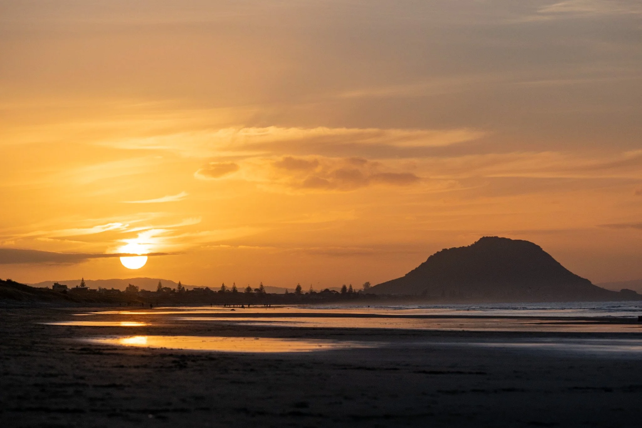 Silhouetted Mount Maunganui at sunset, casting golden reflections on the low tide beach, with an orange sky as the backdrop and the large setting sun in the frame. A peaceful beach landscape captured by ocean photographer Lou Lou B Photo.
