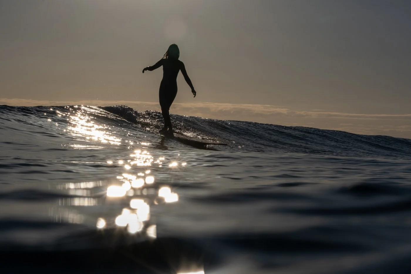 Longboard surfer silhouetted on a wave with sun reflecting across the ocean surface during backlit surf photography