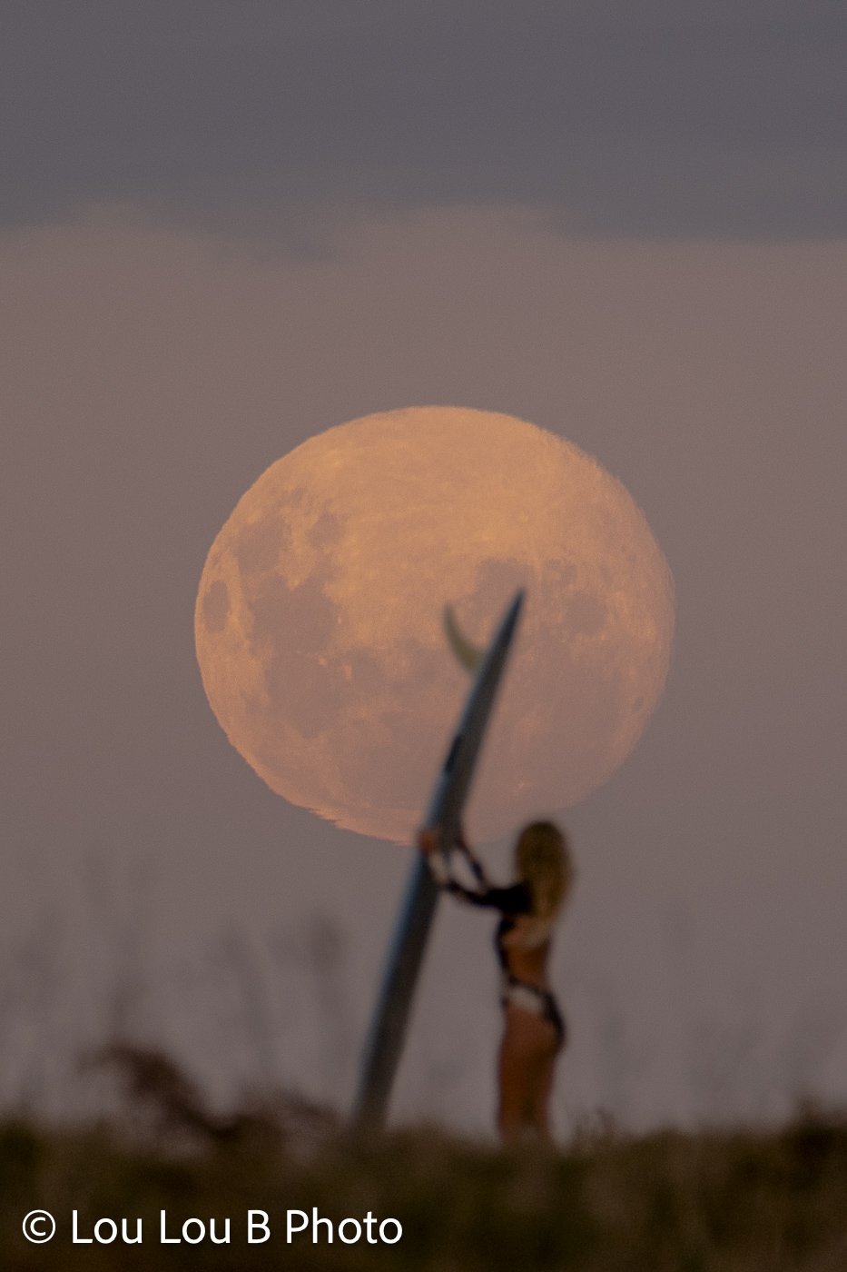 Large golden full moon rising behind a silhouetted person holding a surfboard vertically.