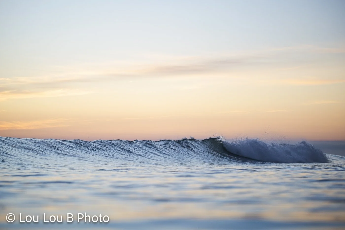 Small ocean wave breaking beneath a soft pastel peach and blue sky.
