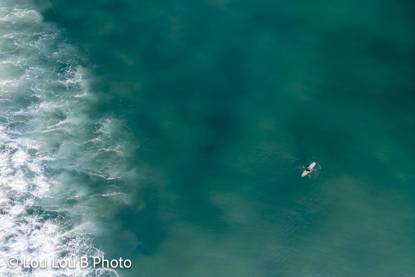 Lone surfer seen from above beside whitewater in deep turquoise ocean.