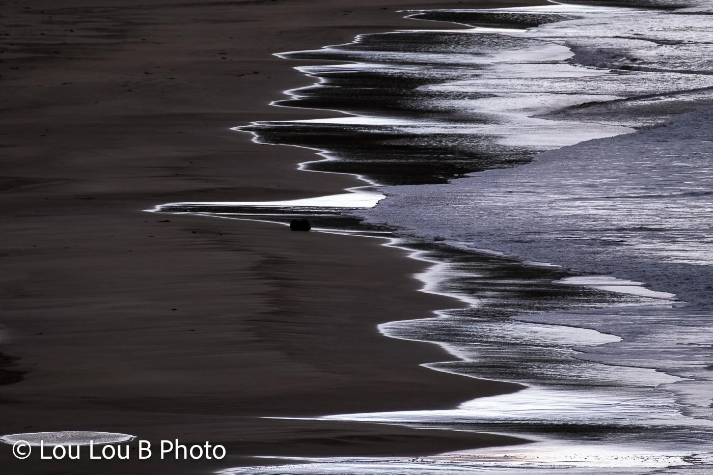 Silvery water tracing the edge of a dark sandy beach in soft coastal light.