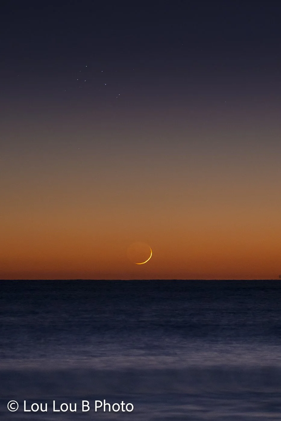 Fine crescent moon and matariki / pleiades / seven sisters star constellation low above the ocean horizon beneath a deep orange and blue twilight sky.