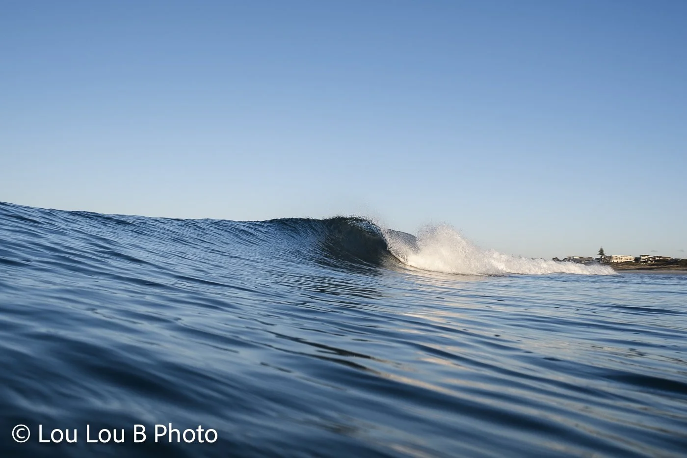 Low water-level view of a clean blue wave breaking with offshore spray beneath a clear sky.