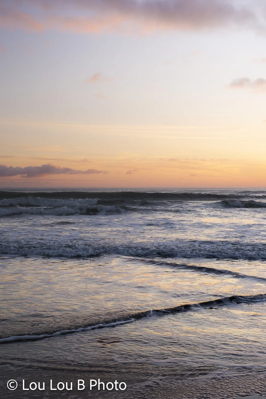 Ocean waves washing onto a reflective beach beneath a soft peach and blue sunset sky.