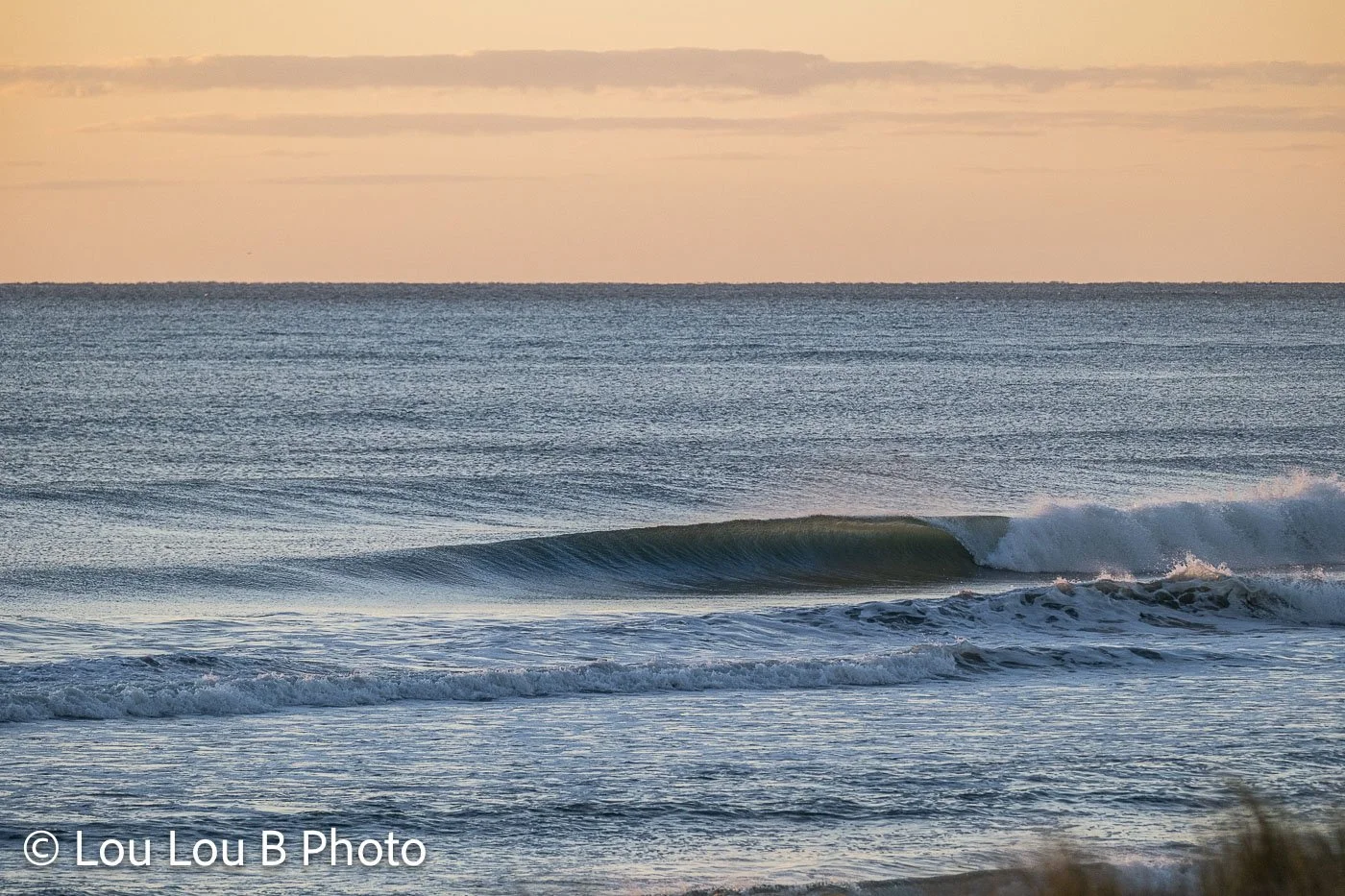 Clean ocean wave peeling across blue water beneath a soft peach evening sky.