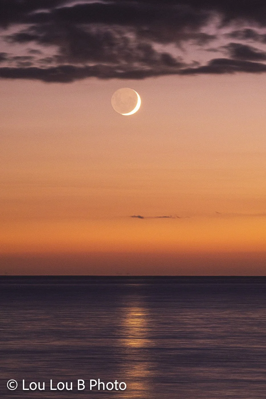 Crescent moon above the ocean at sunset, with warm orange light reflected across the sea.