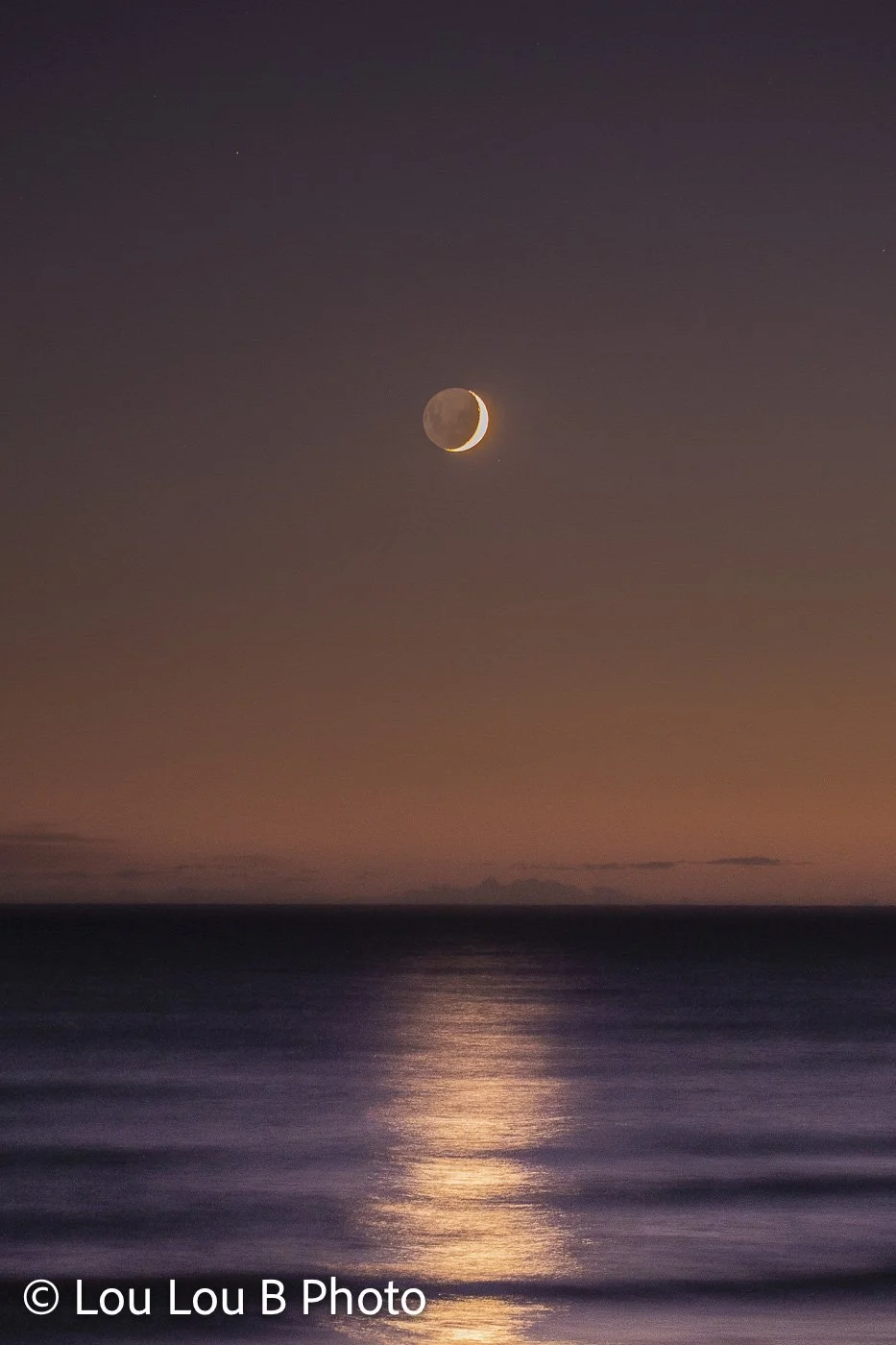 Small crescent moon above the ocean beneath a muted purple and peach evening sky, with moonlight reflected on the sea.