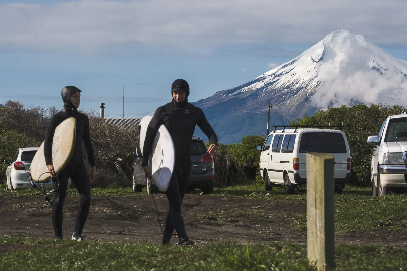 Two surfers carrying surfboards with snow-covered Mount Taranaki rising behind them in Taranaki, New Zealand.