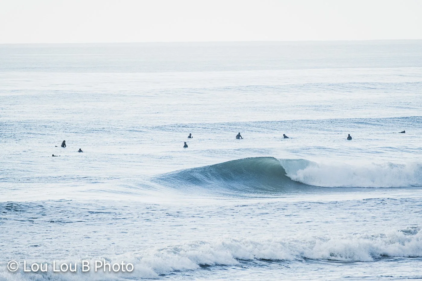 Clean blue-green wave breaking among surfers across a bright, textured ocean.
