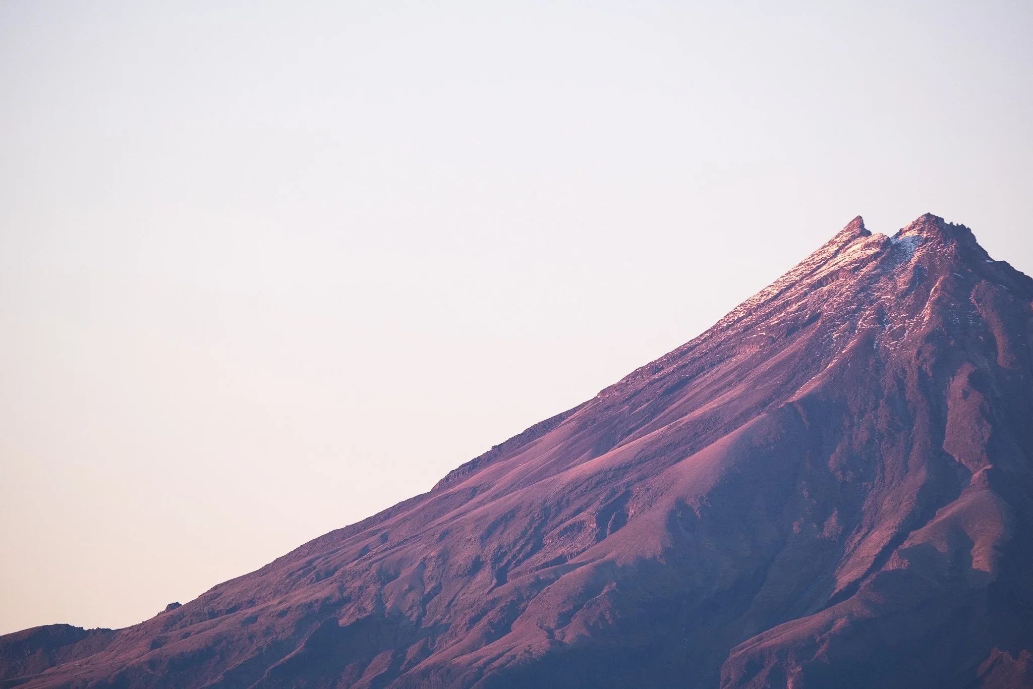 Experience the stunning beauty of New Zealand's landscape in this captivating photograph by Lou Lou B Photo. The iconic Mount Taranaki is captured at sunrise, with its left half, including the peak named Shark's Tooth, appearing in a beautiful pink h