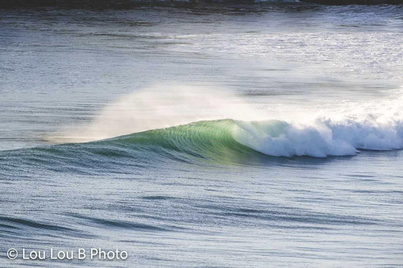 Green-blue wave breaking with offshore spray across the ocean surface.