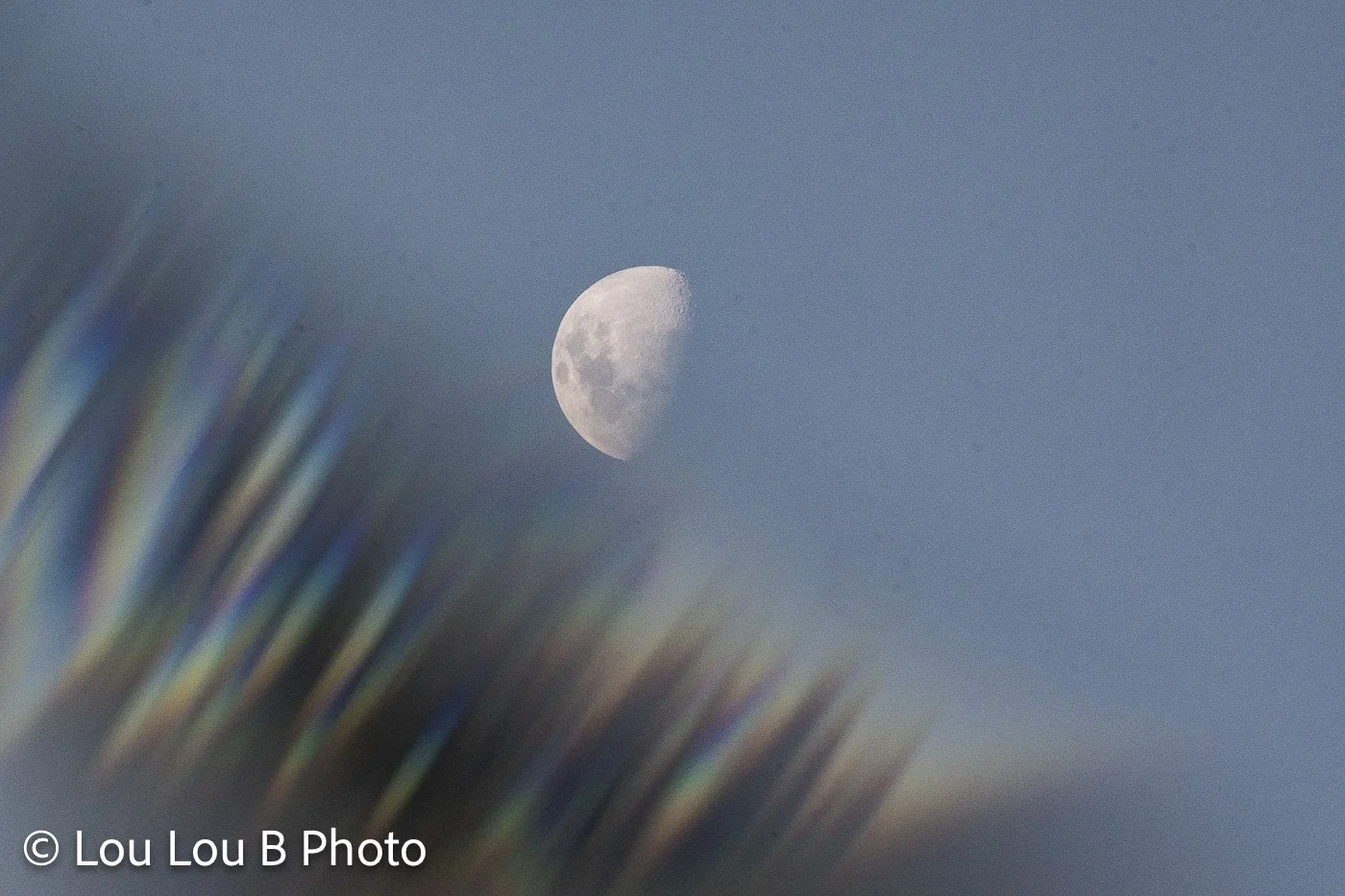Half moon framed by abstract rainbow light and soft blue sky.