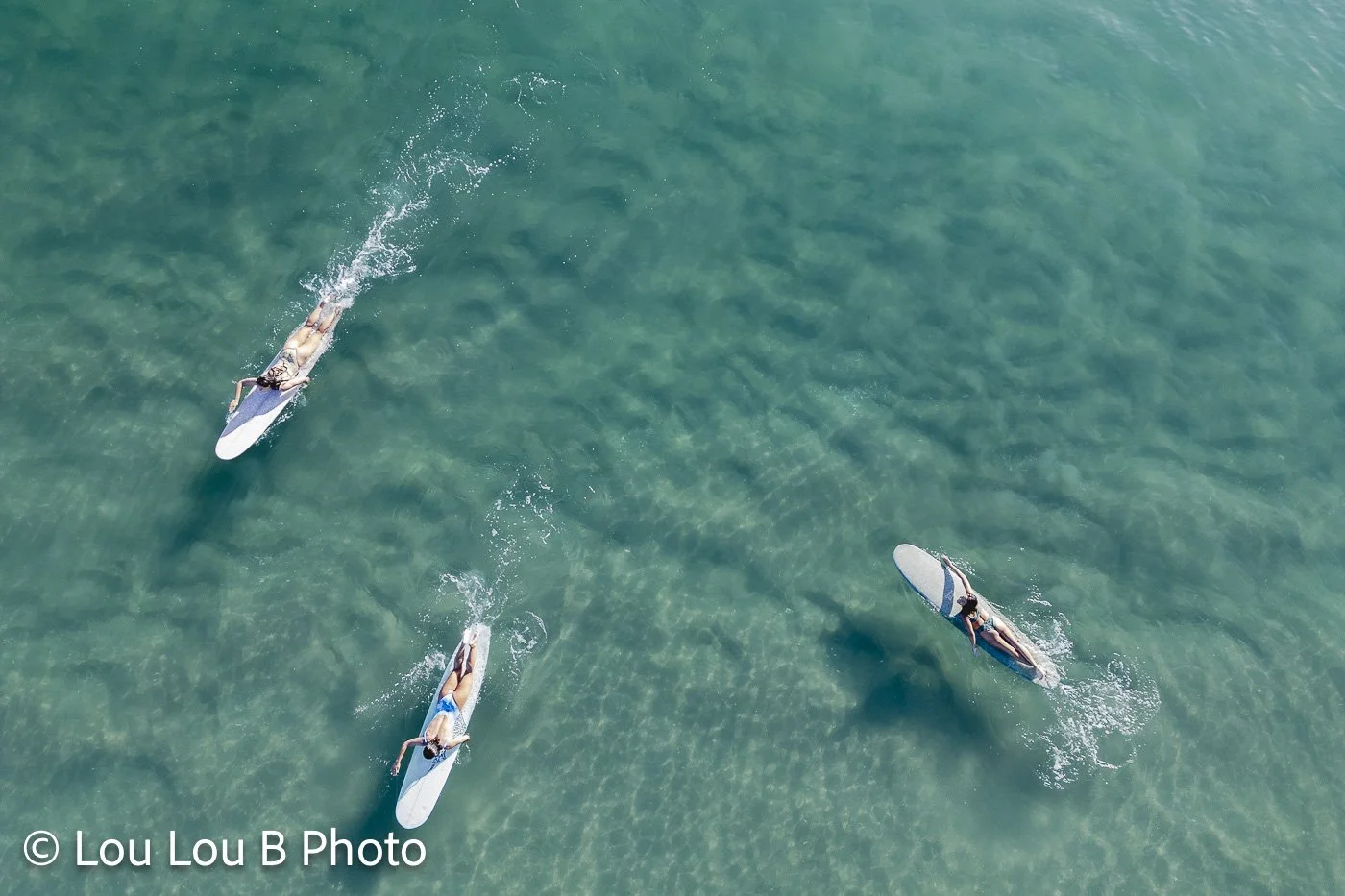 Aerial view of three surfers paddling through clear turquoise ocean water.