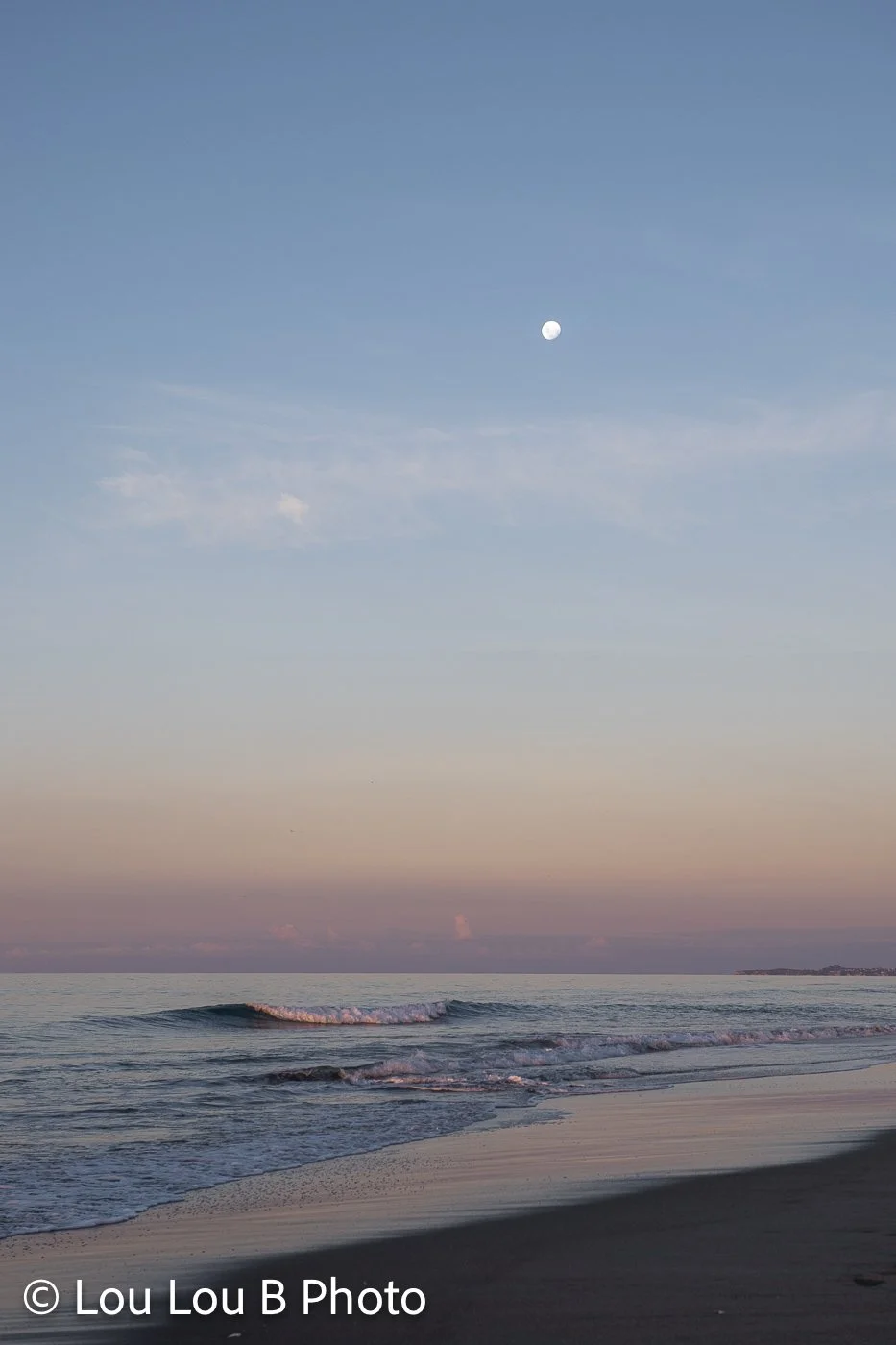 Distant moon above the ocean and beach beneath a soft pastel blue and peach evening sky.