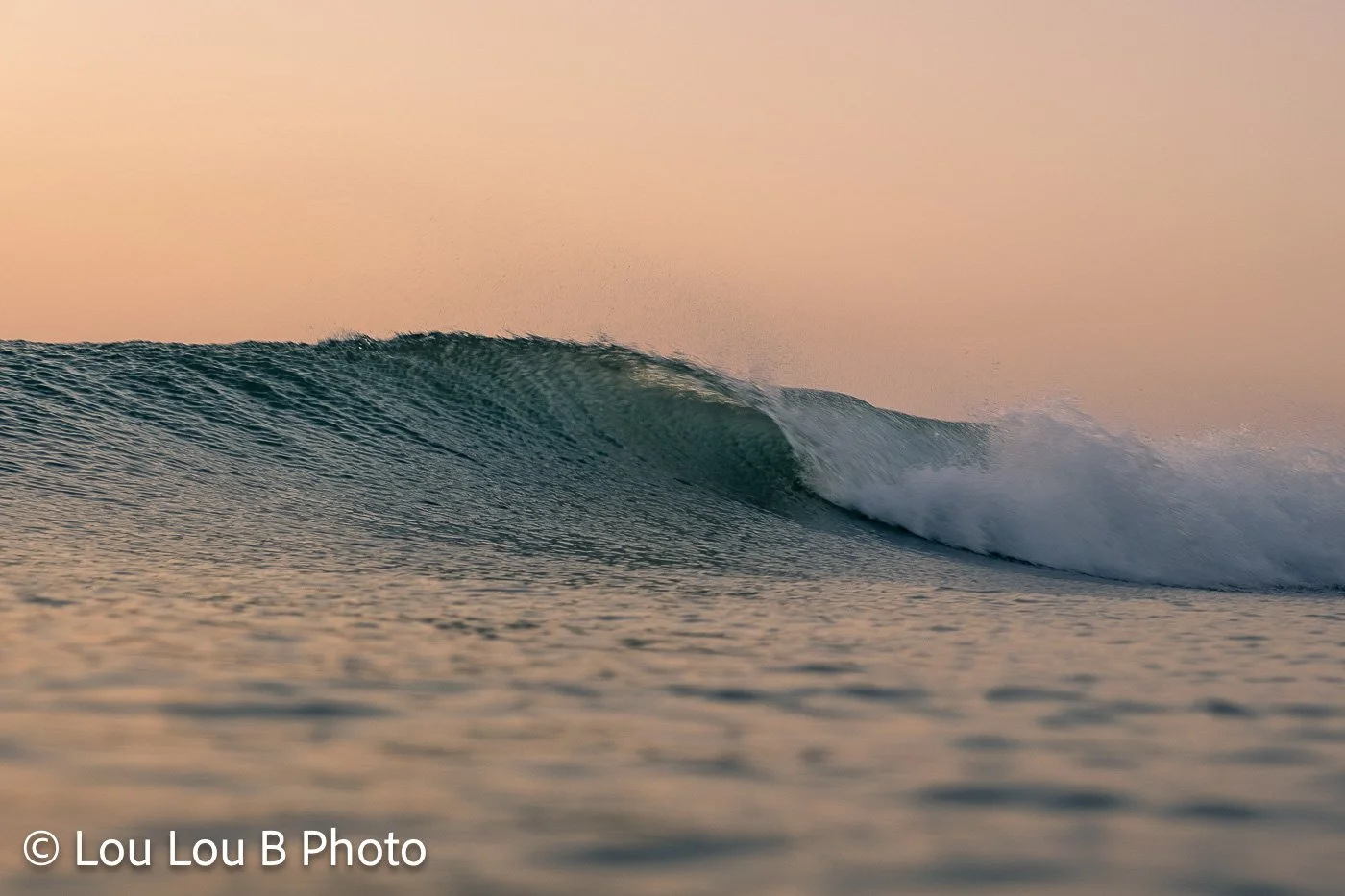 Turquoise wave peeling across the ocean beneath a warm peach sunset sky.