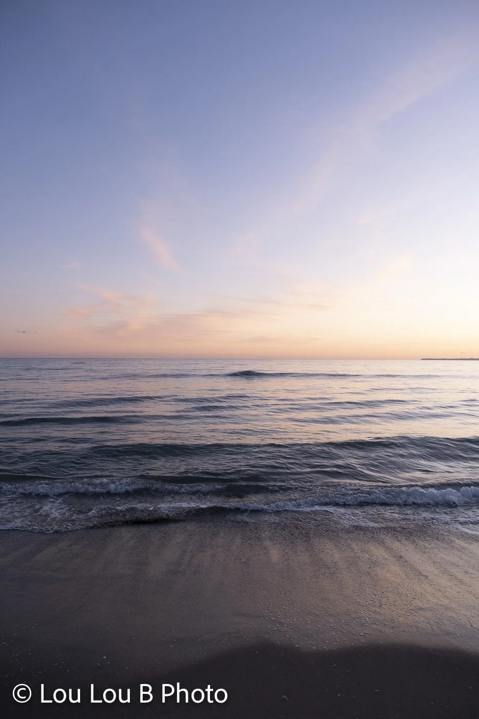 Ocean wave washing onto a dark beach beneath a soft blue, pink and peach sunset sky.