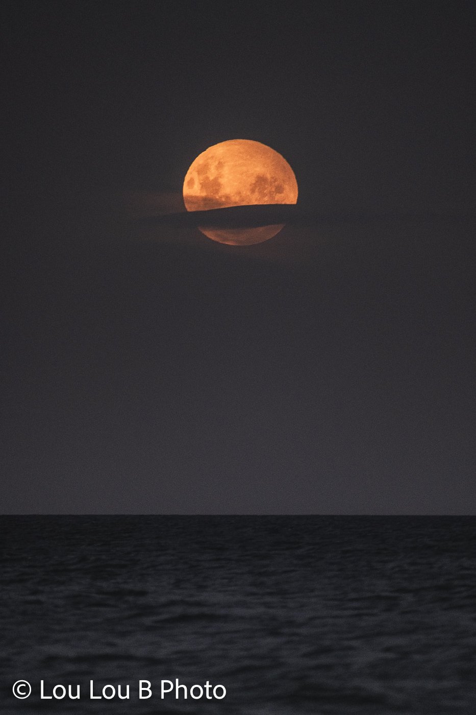 Orange full moon rising above the ocean horizon through a band of cloud.