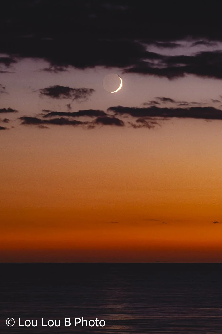 Crescent moon framed by dark clouds above the ocean in a vivid orange and purple sunset sky.