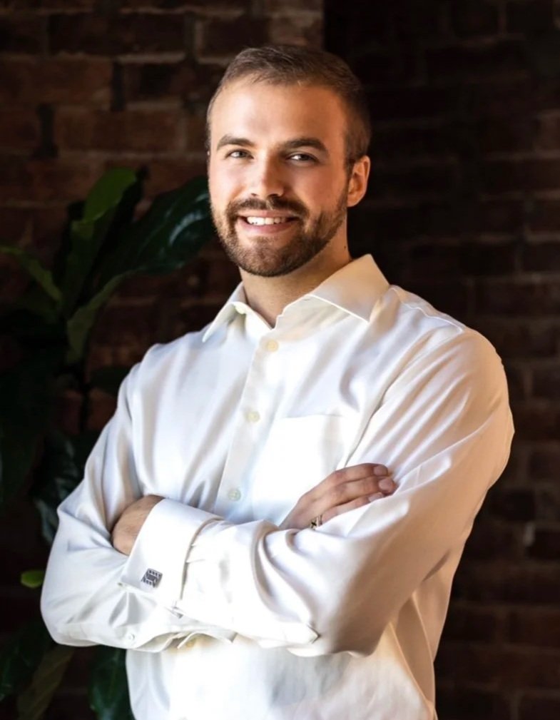 A man with a beard and short hair smiling with arms crossed, standing in front of a brick wall and green plants, wearing a white dress shirt.