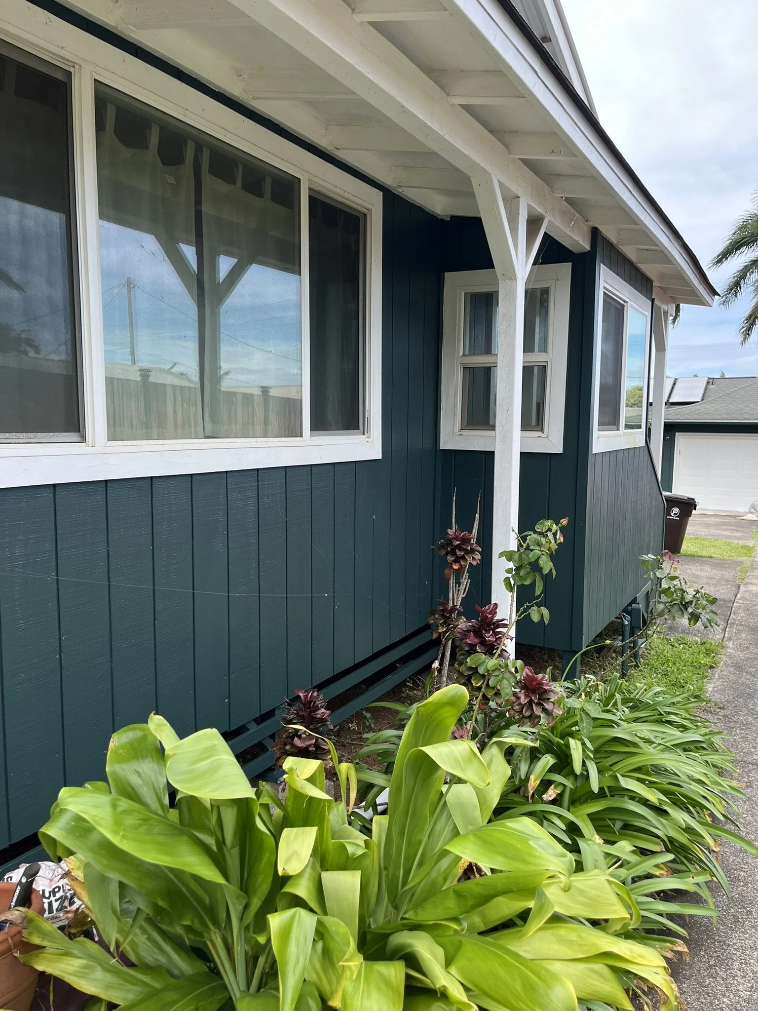 Exterior house painting in Maui - dark green home with white trim and window brackets, surrounded by tropical landscaping, by Maui Home Refinishing.
