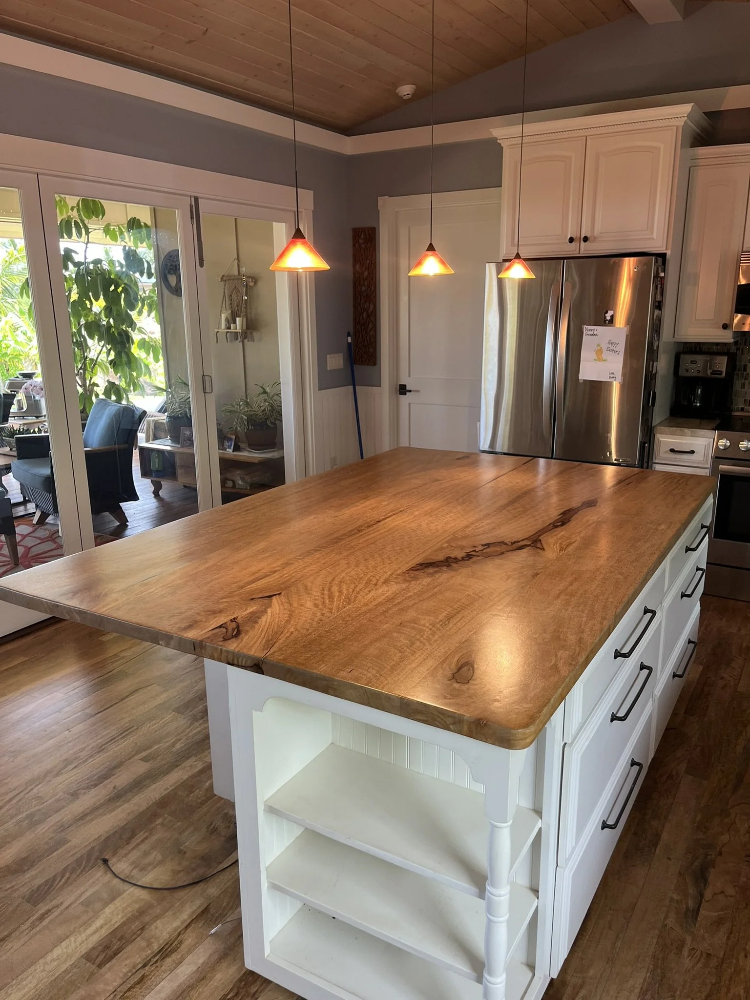 Cabinet refinishing in a Maui home kitchen - wood island with white base cabinets, pendant lighting, and stainless steel refrigerator, by Maui Home Refinishing.