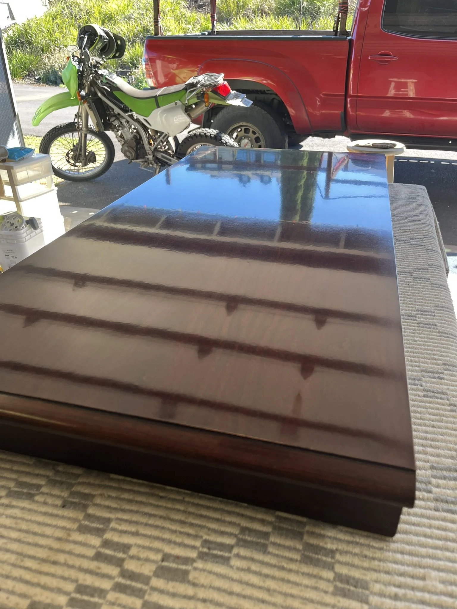 Wood table refinishing in a Maui home - view from inside showing a reflective wood table finish, with a truck and motorcycle parked outside.