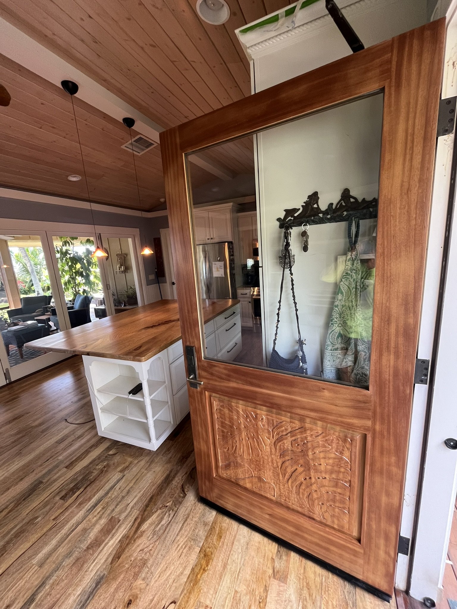 View of a kitchen with a wooden ceiling, pendant lights, and a sliding glass door leading to an outdoor patio area with trees. Inside, there is a wooden countertop, white cabinetry, and wall decor.