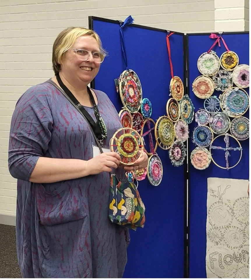 Image shows a woman holding a piece of textile art and in the background is a larger textile artwork.