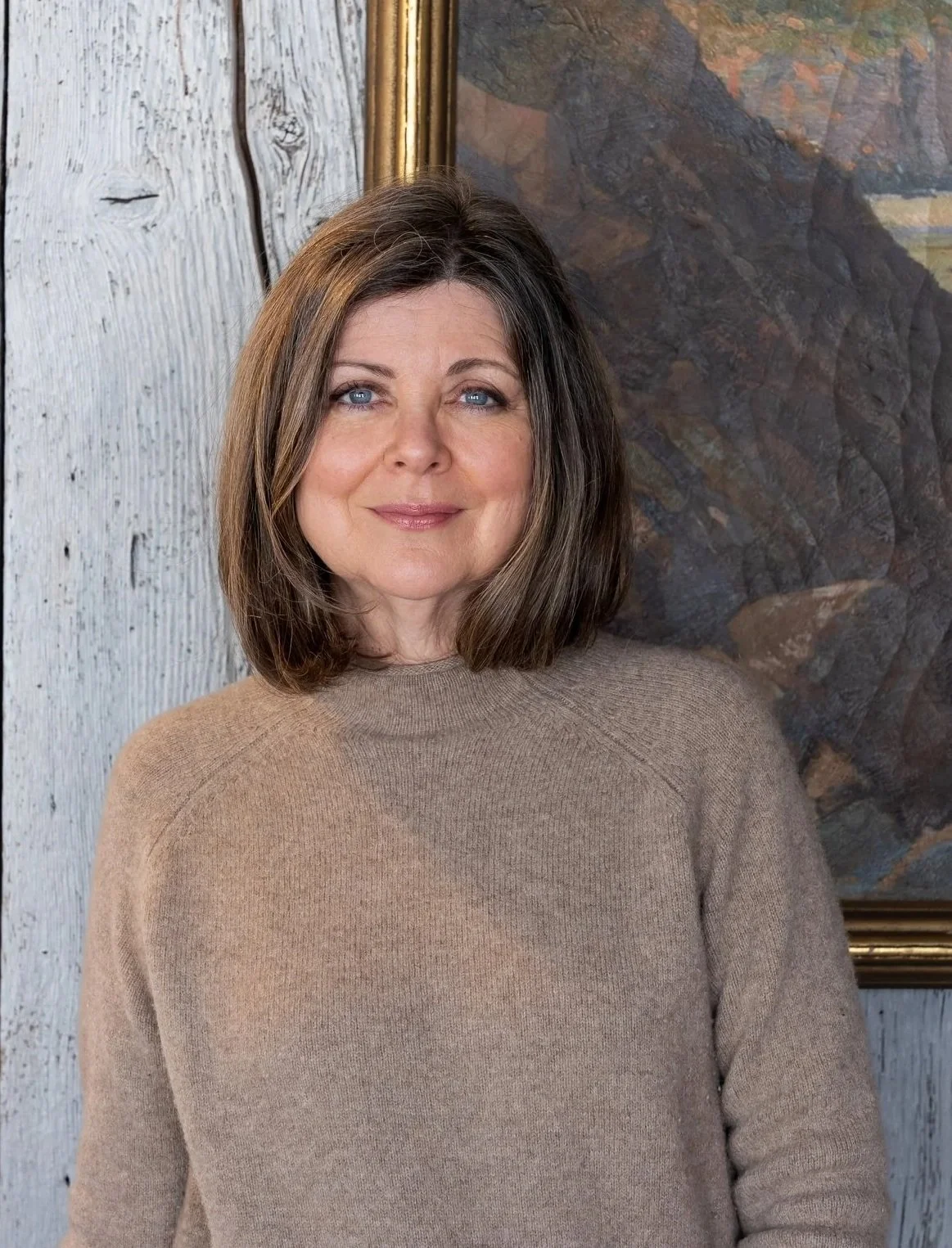 A woman with shoulder-length brown hair and blue eyes smiling, wearing a beige sweater, standing in front of a framed painting and a rustic wooden wall.