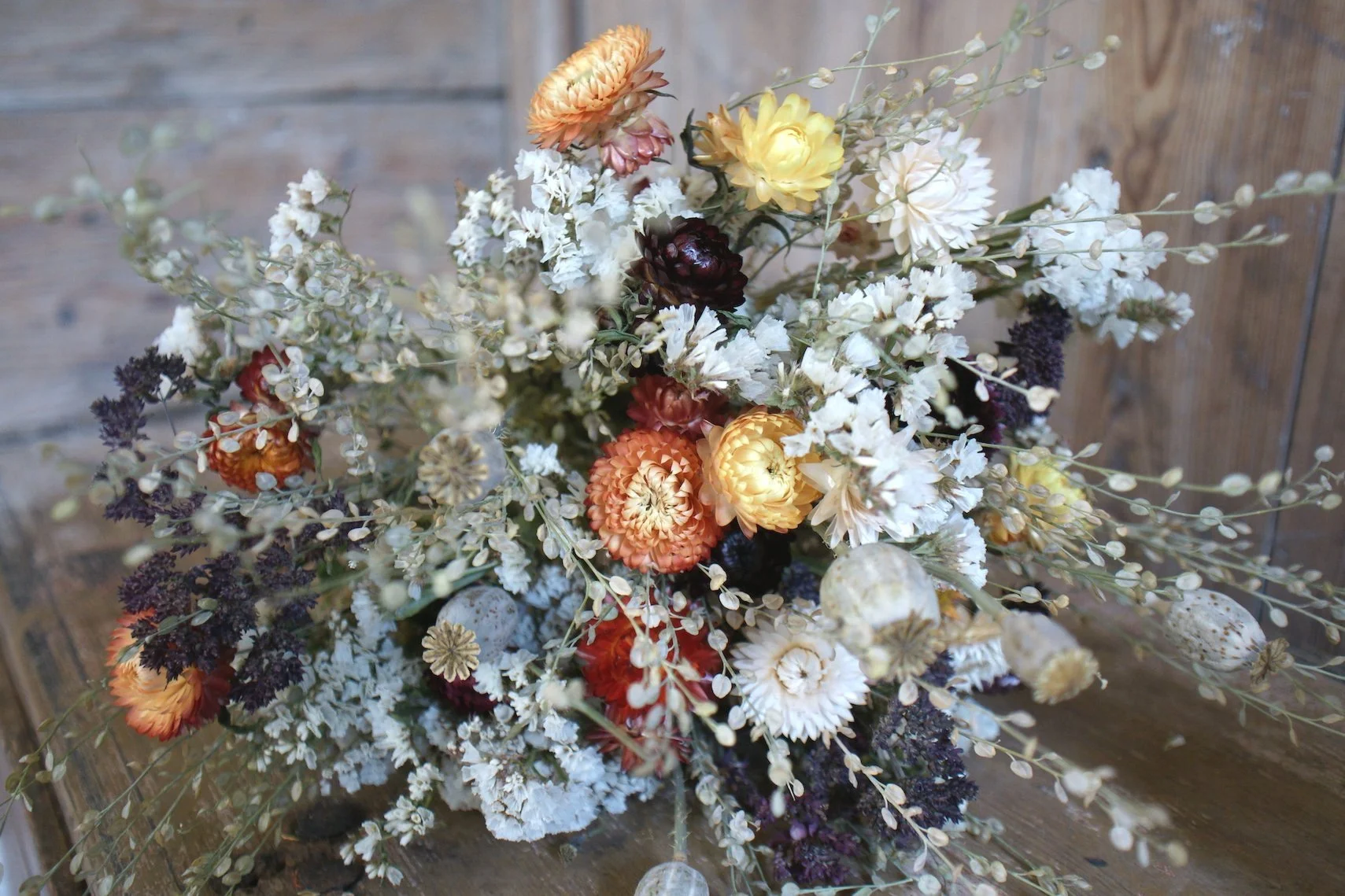 En bukett med tørkede blomster, inkludert flere blomster i oransje, gule og hvite toner, omgitt av tørre blader og frøkapsler, plassert foran en trebakgrunn.