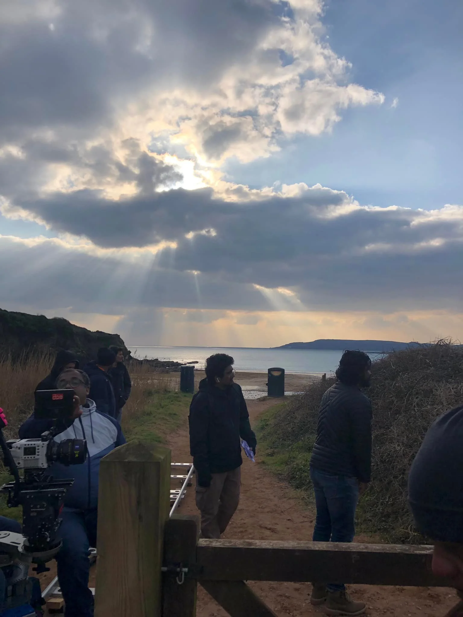 People gathered outdoors near a beach on a cloudy day with rays of sunlight breaking through the clouds.
