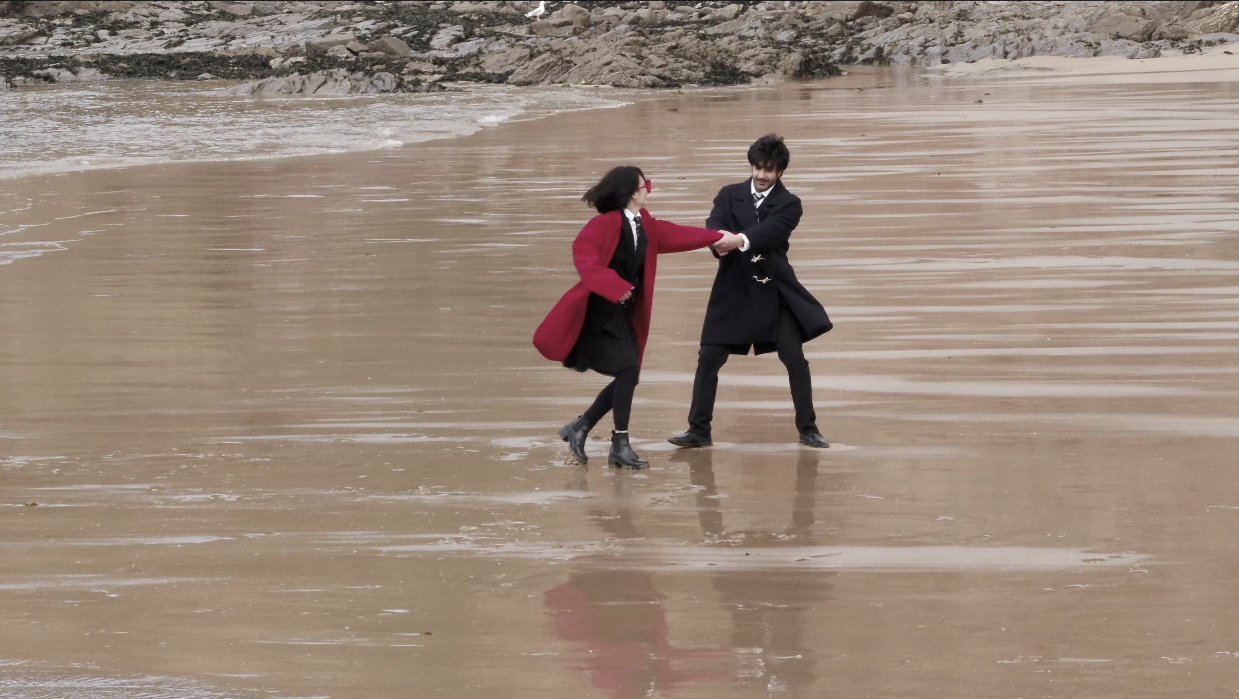 Two people dressed in black and red coats fighting on a beach with wet sand, with ocean waves and rocks in the background.