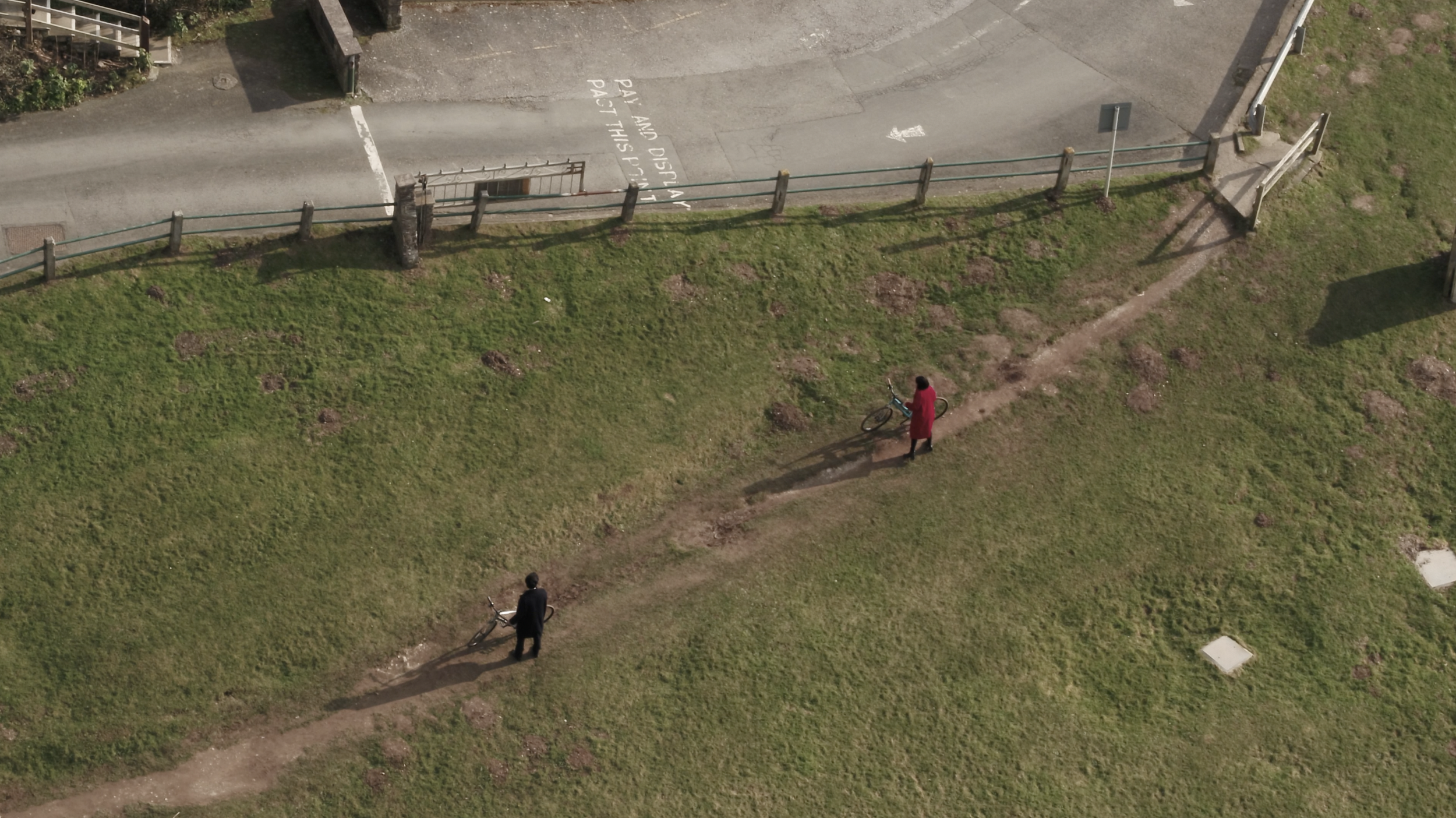 An aerial view of a grassy park area with a dirt path. Two people are walking their bicycles along the path, one in a black outfit and the other in a red coat. There is a street with a fence and some parked or passage vehicles visible at the top of t
