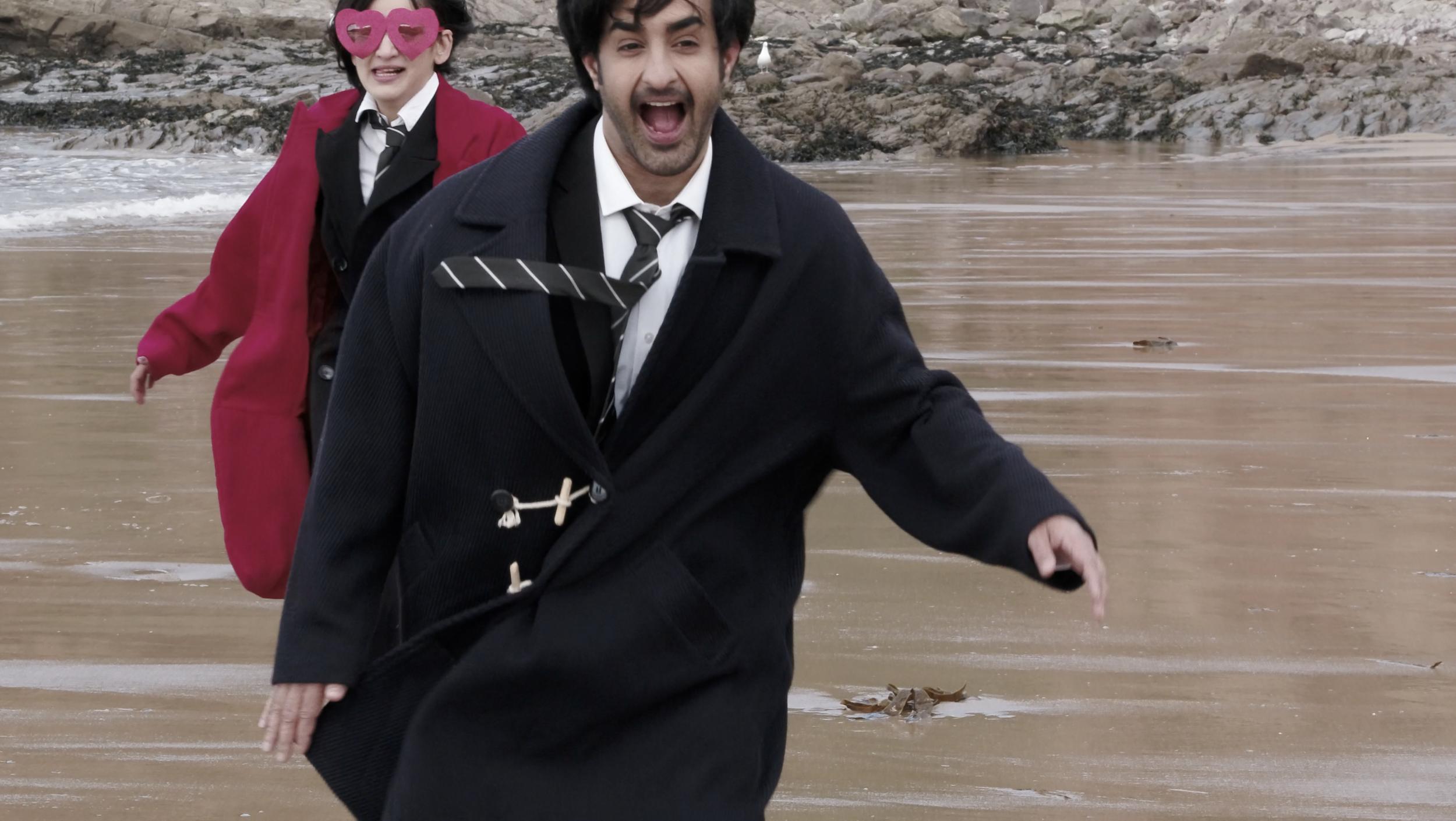 Two people dressed in black coats and ties running on a sandy beach, with the man in front and a woman with pink heart-shaped sunglasses behind him, near rocks and water.